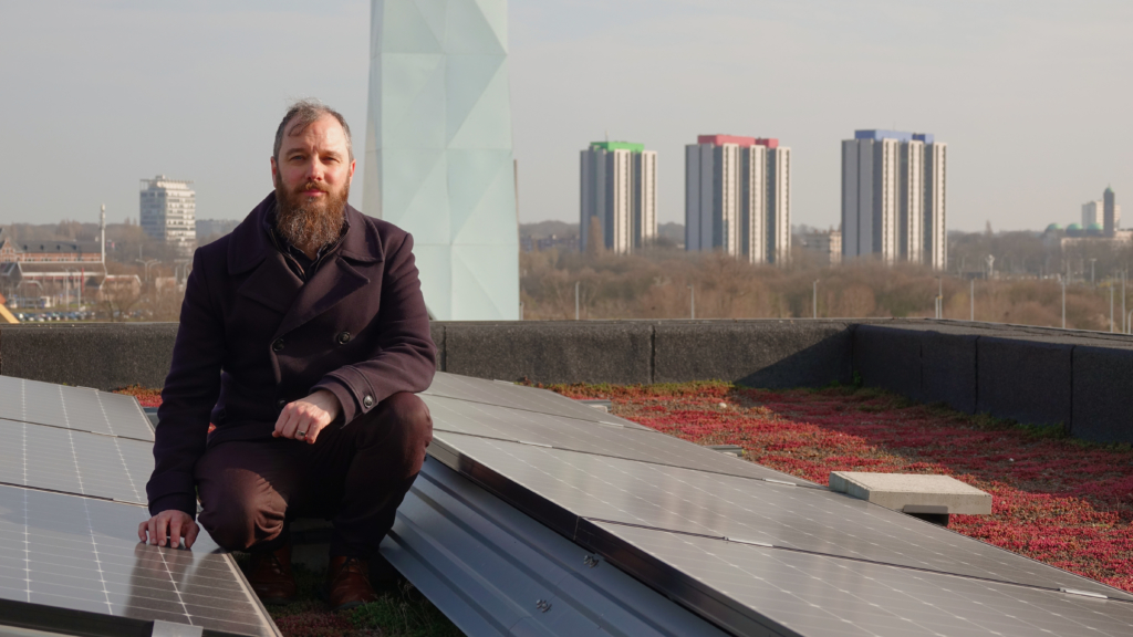 Jan Wouters op een dak van één van onze gebouwen op Nieuw Zuid. Op het dak staan zonnepanelen. Op de achtergrond staat de verwarmingstoren die de wijk van warm water voorziet.